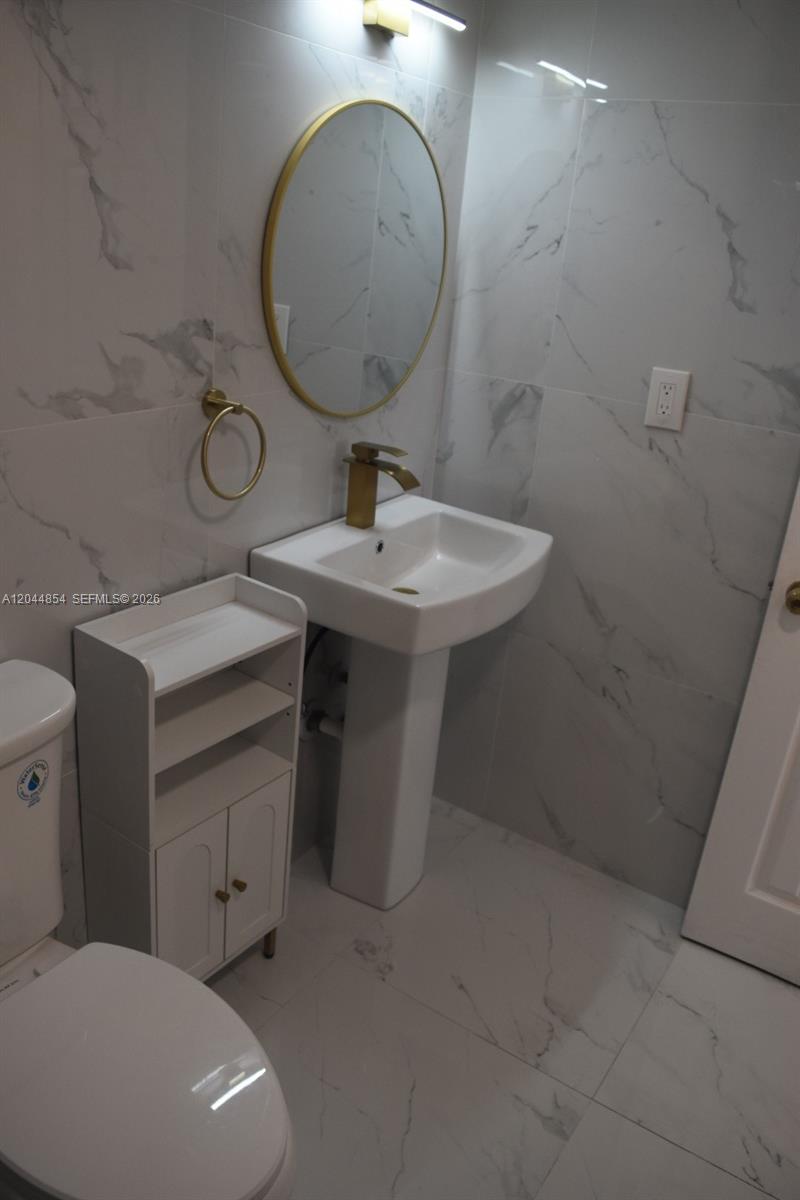 A bathroom features a pedestal sink, a round gold-framed mirror, and white marble-patterned tile — 666 W 81st St # 311, Hialeah FL 33014, HIALEAH, FL