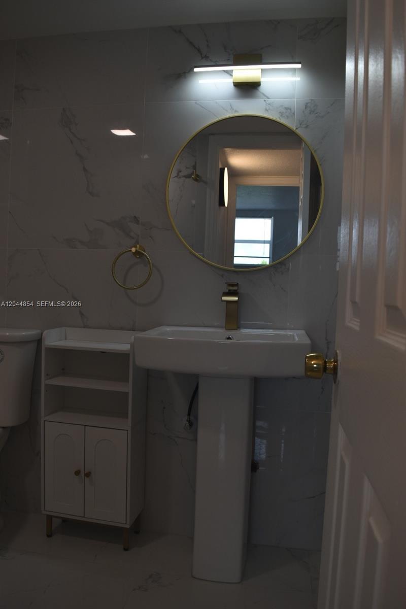 A bathroom features a pedestal sink, a round gold-framed mirror, and marble-patterned wall tiles — 666 W 81st St # 311, Hialeah FL 33014, HIALEAH, FL