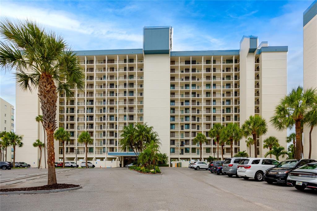 A multi-story apartment building with balconies stands behind a paved parking lot with palm trees — 7100 SUNSET WAY UNIT 810, ST PETE BEACH, FL