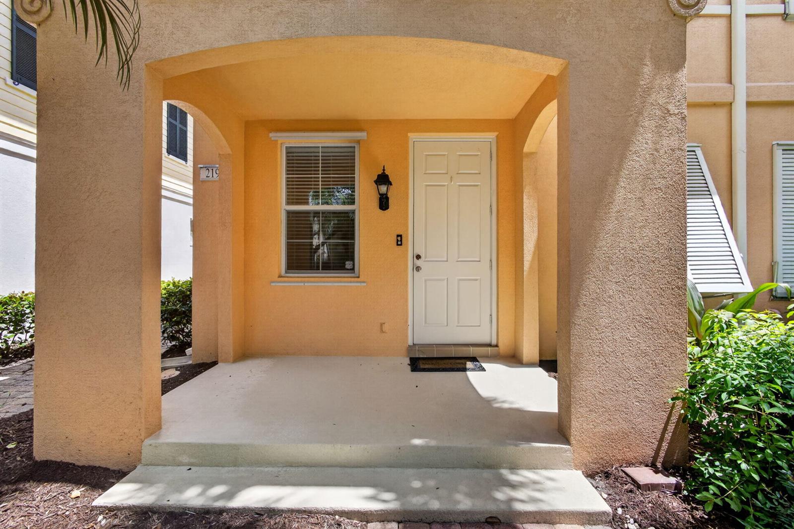 A covered entryway features a beige stucco exterior, a white front door, and a window — 219 Soriano Drive, Jupiter FL 33458, JUPITER, FL