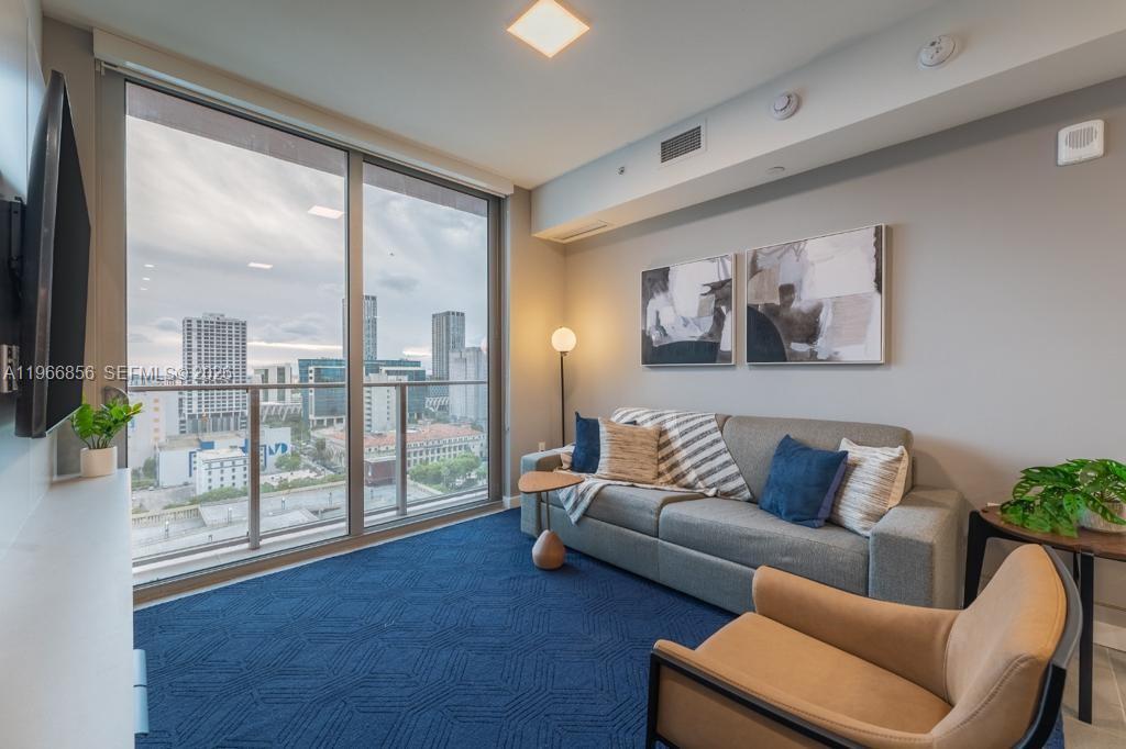 A living room features a gray sofa, blue carpet, and floor-to-ceiling windows overlooking a city — 227 NE 2nd St # 1905, Miami FL 33132, MIAMI, FL