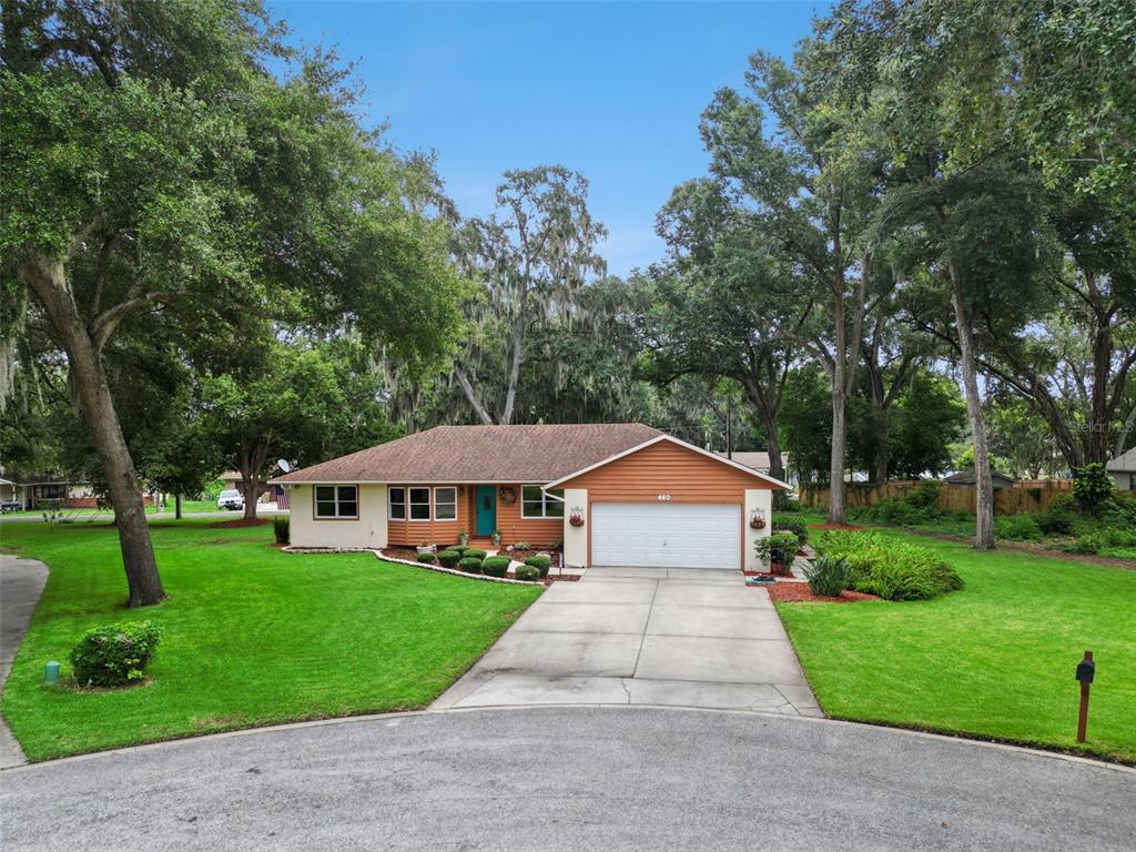 A single-story house with tan siding, a two-car garage, and a large lawn with trees — 480 LAKE FOREST CIRCLE, MOUNT DORA, FL