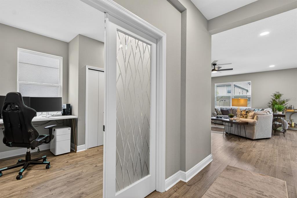 A home office with a glass-paneled door opens into a living room with wood flooring — 7241 NOTCHED PINE BEND, WESLEY CHAPEL, FL