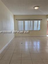 An empty room features light-colored tile flooring, cream-colored walls, and a large window with blinds — 15139 NE 6th Ave # 3, Miami FL 33162, MIAMI, FL