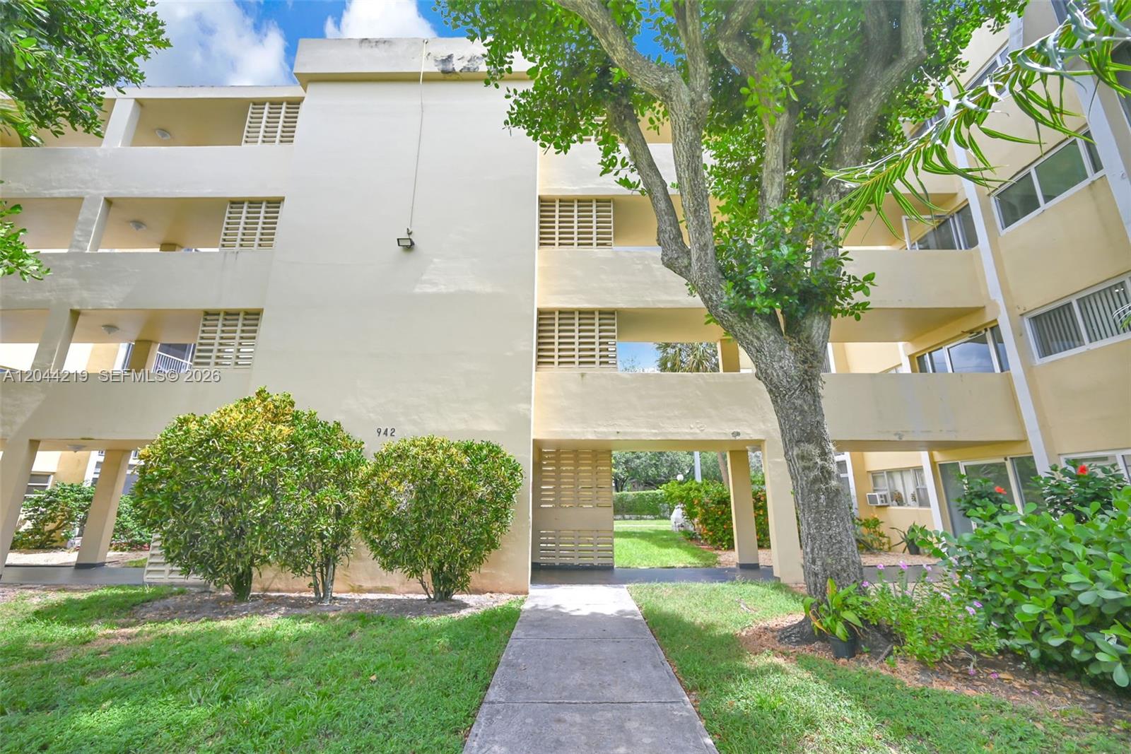 A concrete walkway leads to the entrance of a multi-story apartment building with balconies — 942 NE 199th St # 4E, Miami FL 33179, MIAMI, FL