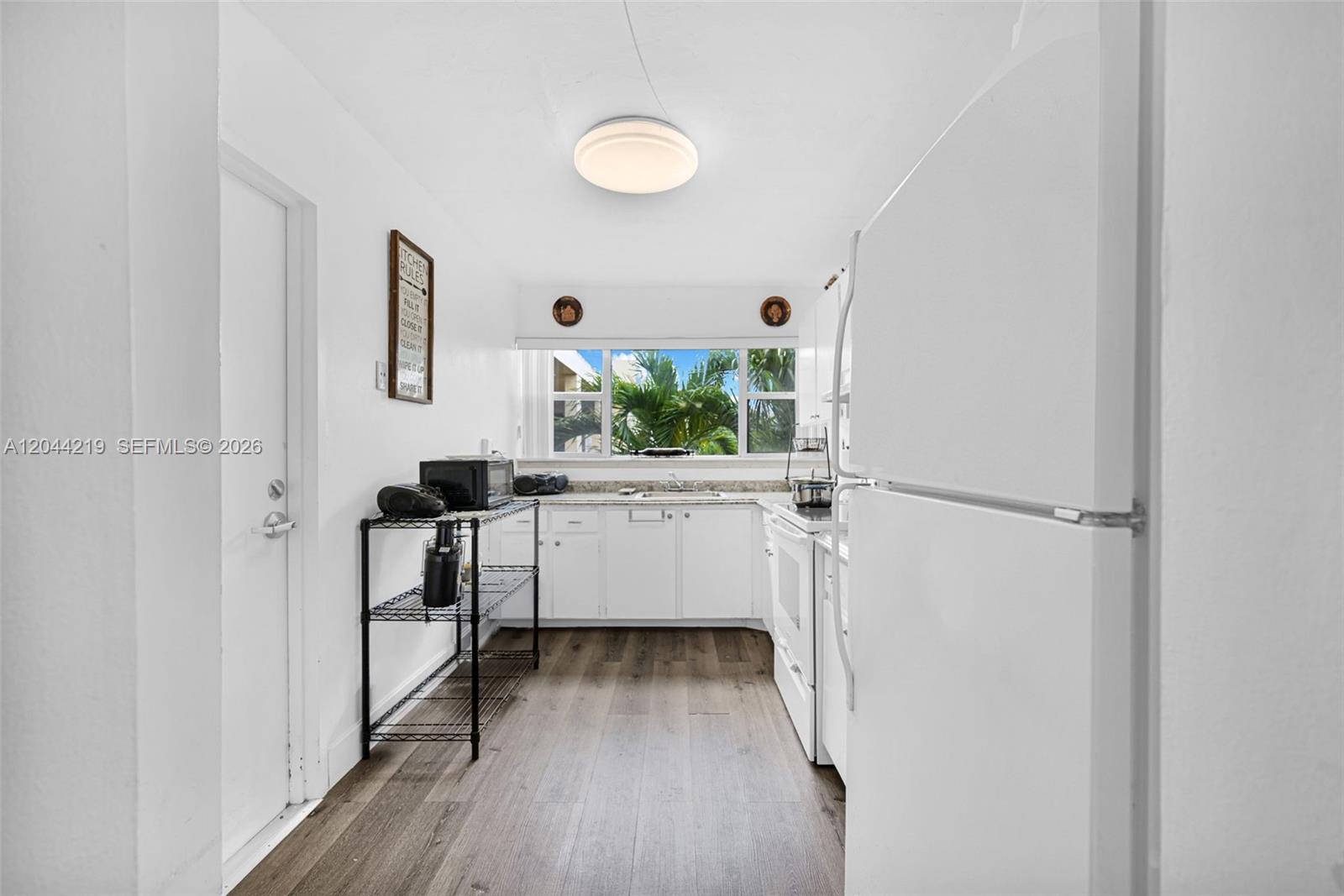 A white kitchen features wood-look flooring, a wire shelving unit, and a window with palm trees — 942 NE 199th St # 4E, Miami FL 33179, MIAMI, FL