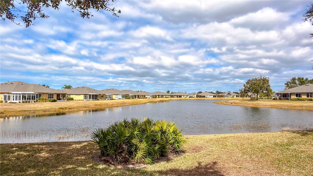 A pond sits in front of several suburban houses under a cloudy blue sky — 2069 SIFIELD GREENS WAY UNIT 102, SUN CITY CENTER, FL