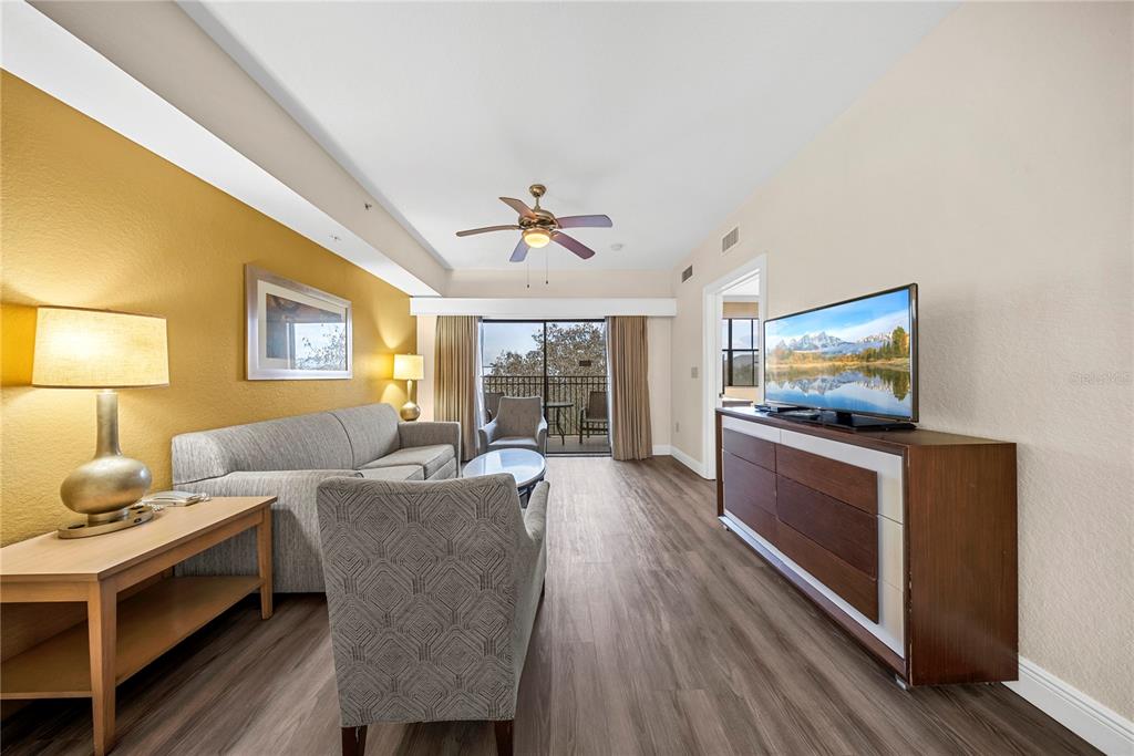 A living room features a grey sofa, patterned armchair, wood-look flooring, and a balcony view — 12527 FLORIDAYS RESORT DRIVE UNIT 402E, ORLANDO, FL