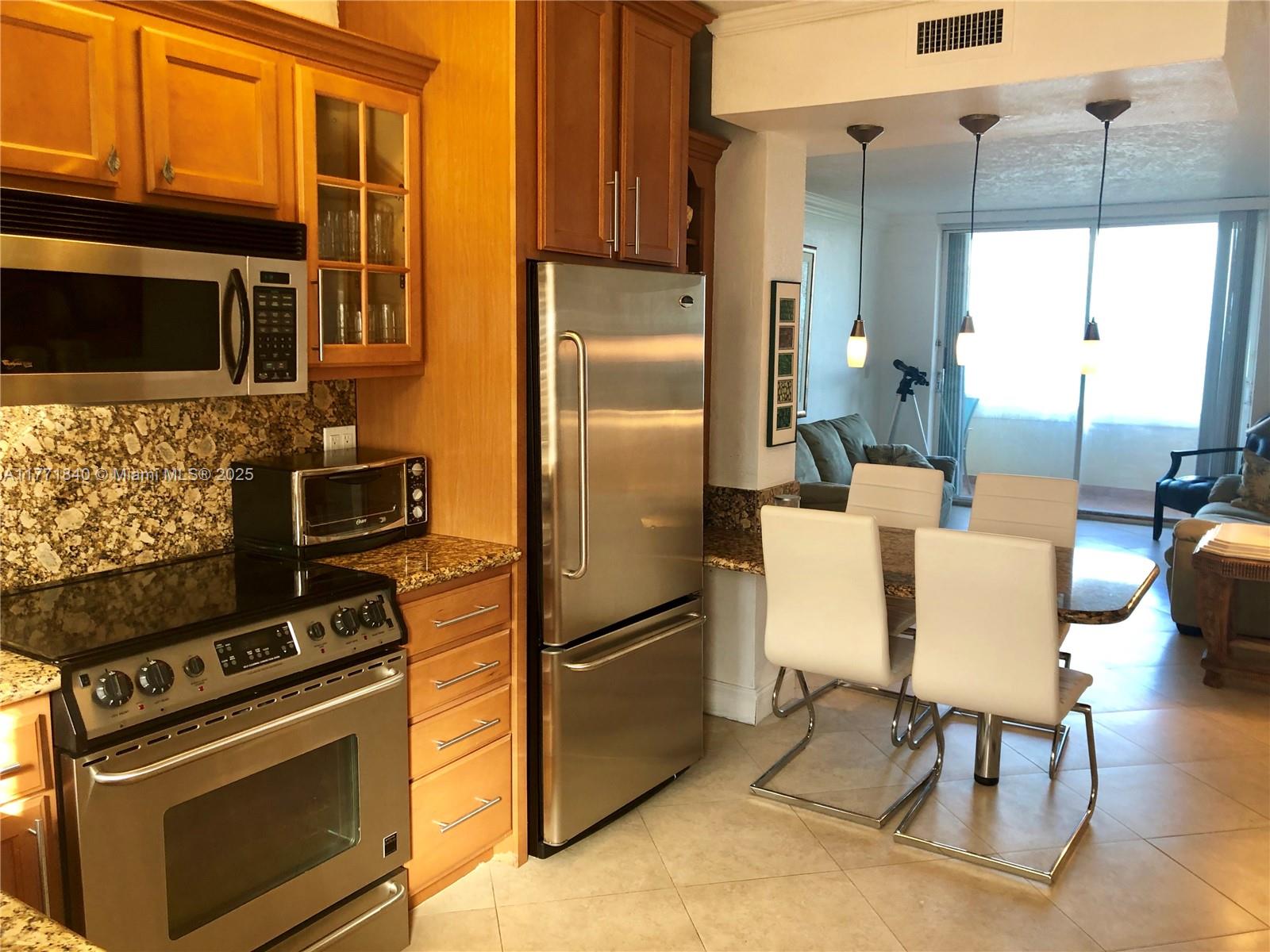 A kitchen with stainless steel appliances and wood cabinets opens into a dining area — 1936 S Ocean Dr # 24C, Hallandale Beach FL 33009, HALLANDALE BEACH, FL