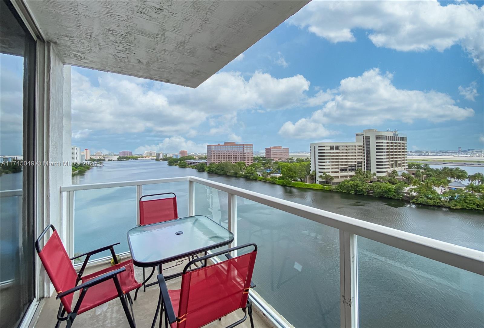 A balcony with a glass table and three red chairs overlooks a river and city buildings — 5091 NW 7th St # 1001, Miami FL 33126, MIAMI, FL