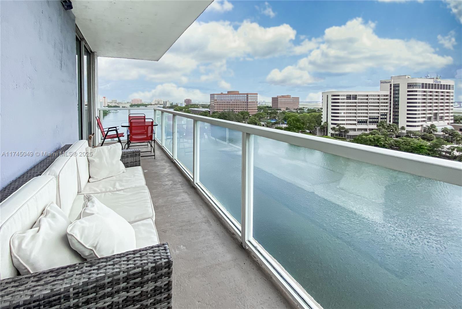 A balcony features a wicker sofa, red chairs, and a glass railing overlooking a waterway — 5091 NW 7th St # 1001, Miami FL 33126, MIAMI, FL
