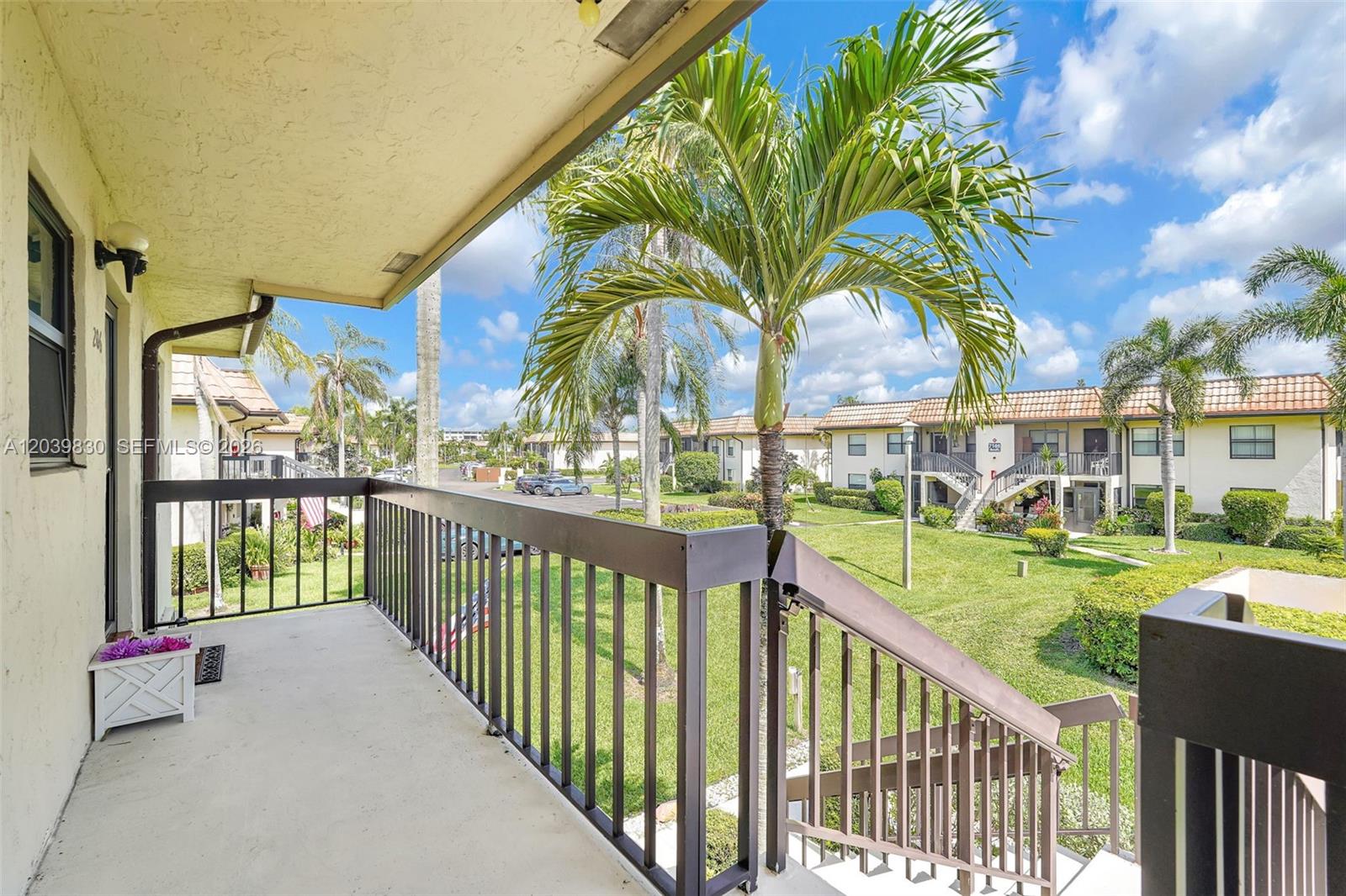 A balcony with a metal railing overlooks a grassy courtyard with palm trees and buildings — 7142 Golf Colony Ct # 206, Lake Worth FL 33467, LAKE WORTH, FL