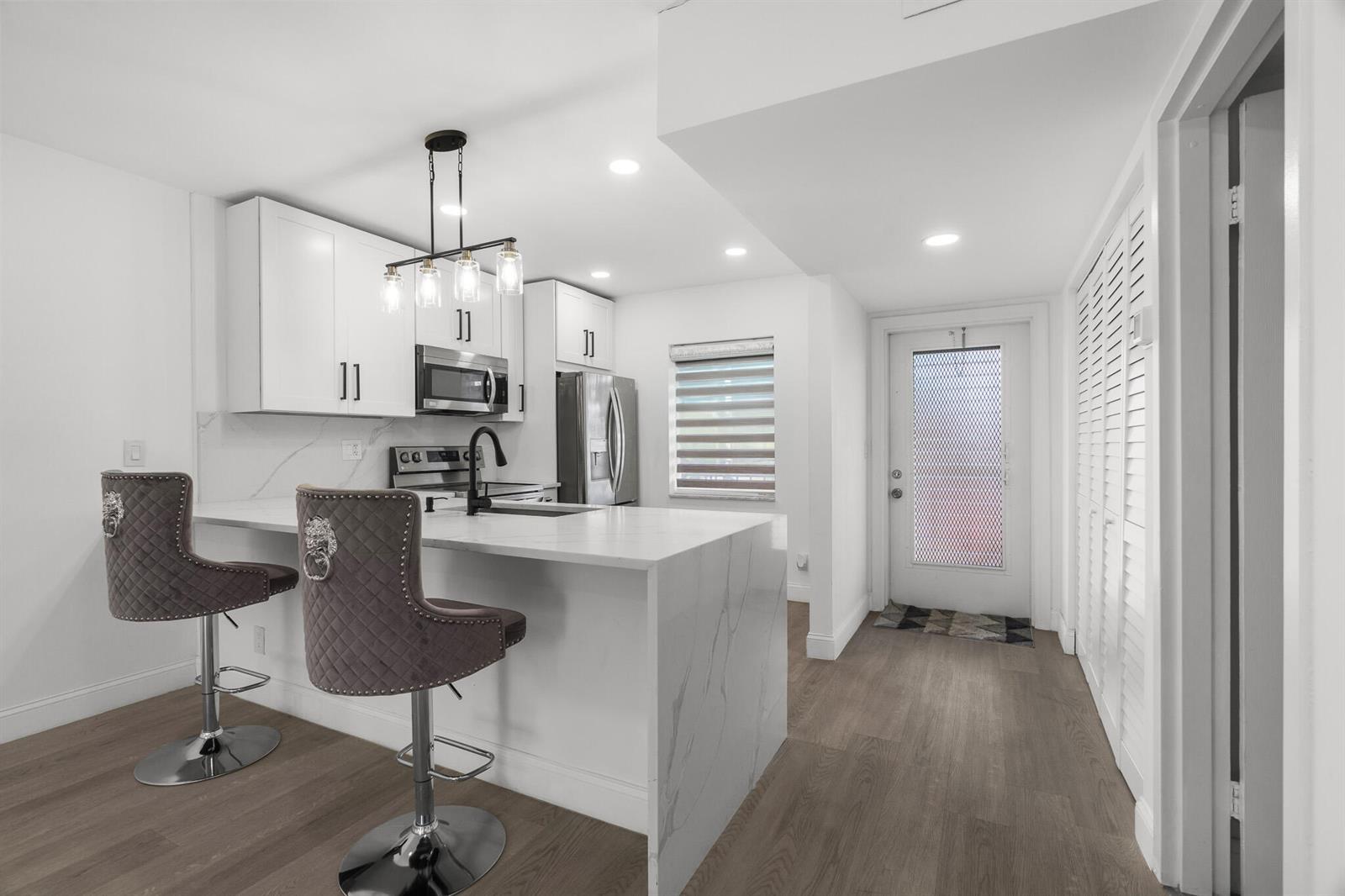 A modern kitchen features white cabinets, a marble-topped island with two barstools, and wood flooring — 5 Abbey Lane # 207, Delray Beach FL 33446, DELRAY BEACH, FL