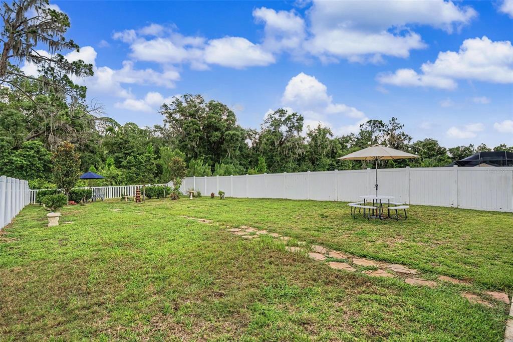 A grassy backyard features a stone path, a white vinyl fence, and a patio umbrella — 13411 WILLOW BLUESTAR LOOP, RIVERVIEW, FL