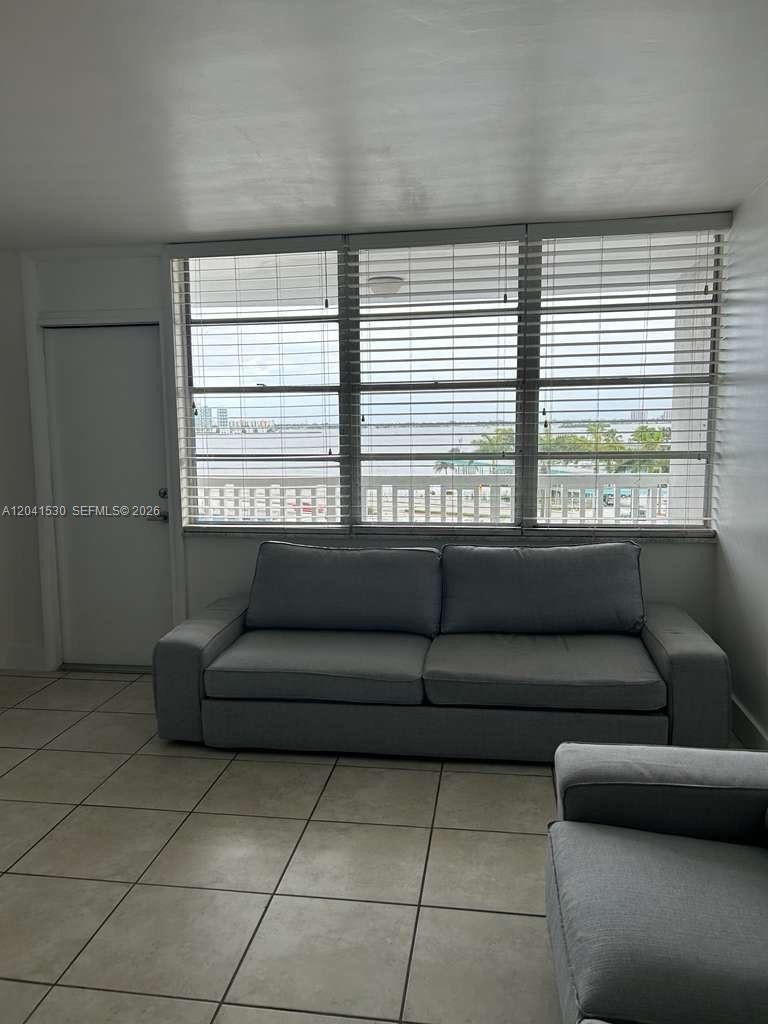 A living room features a gray sofa, tiled floors, and large windows overlooking the water — 2016 Bay Dr # 602, Miami Beach FL 33141, MIAMI BEACH, FL