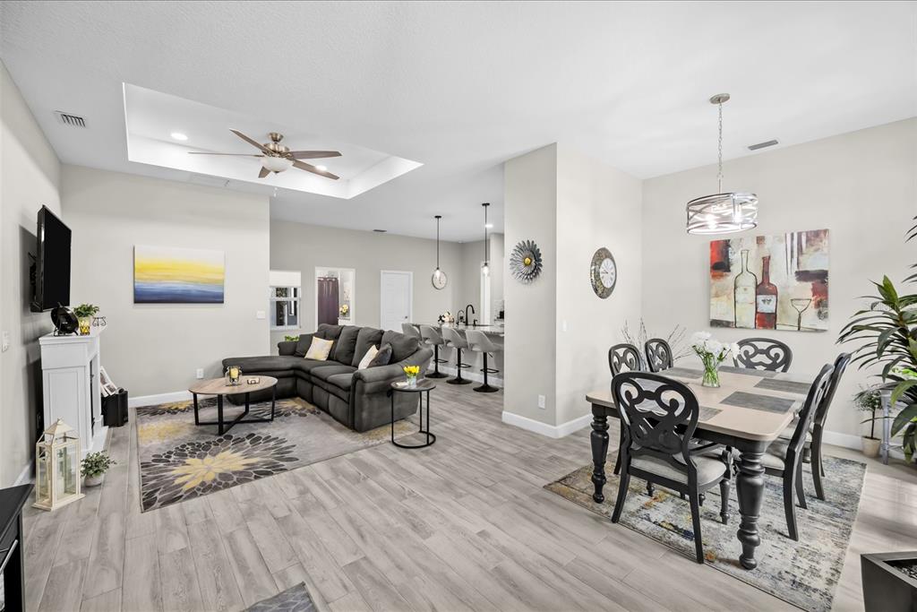 An open-concept living and dining area features gray wood-look flooring, a sectional sofa, and lighting — 1980 SW AMERICANA STREET, PORT ST LUCIE, FL