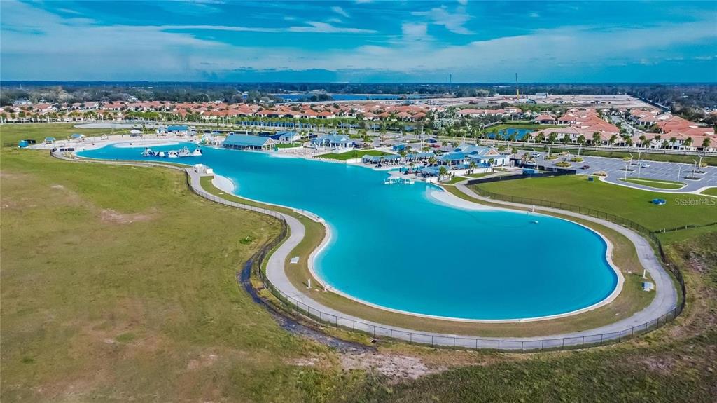 An aerial view shows a large, bright blue swimming lagoon surrounded by a paved walkway — 17029 BLISTER WING DRIVE, WIMAUMA, FL