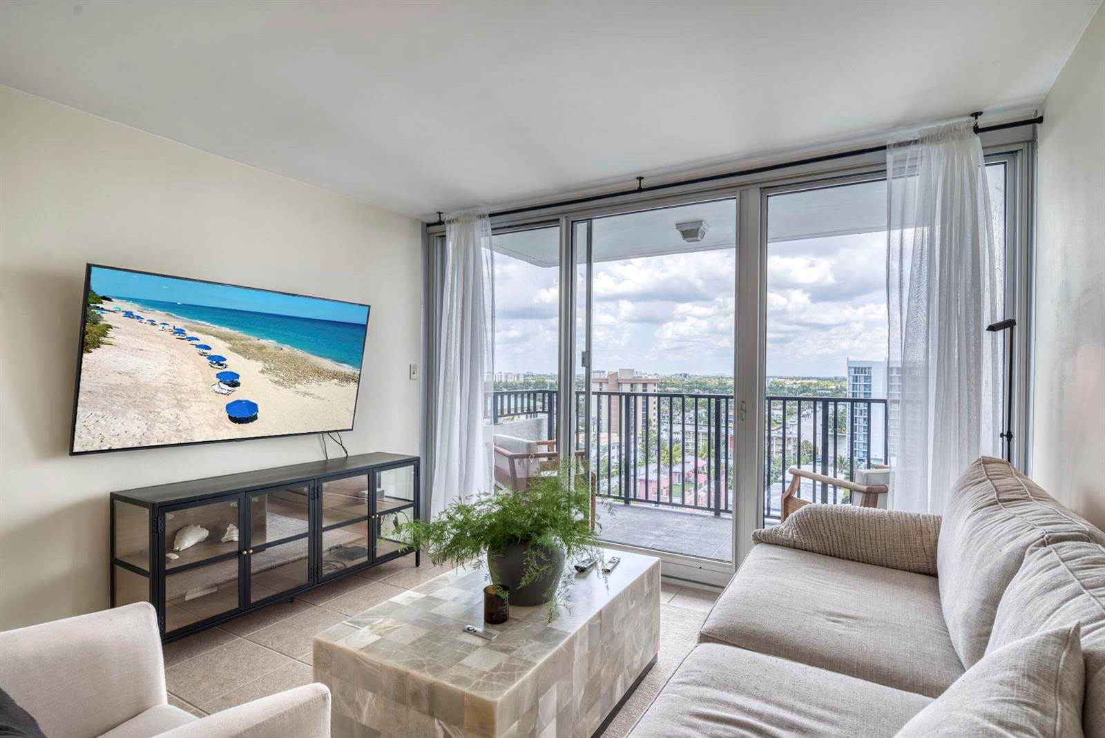 A living room features a wall-mounted television, a stone coffee table, and a balcony view — 525 N Ocean Boulevard # 1215, Pompano Beach FL 33062, POMPANO BEACH, FL