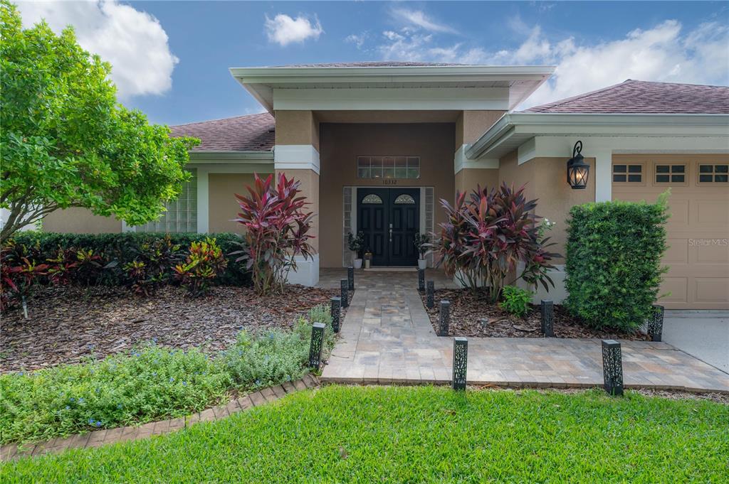 A paved walkway leads to the double front doors of a tan house with landscaping — 10332 ABBOTSFORD DRIVE, TAMPA, FL