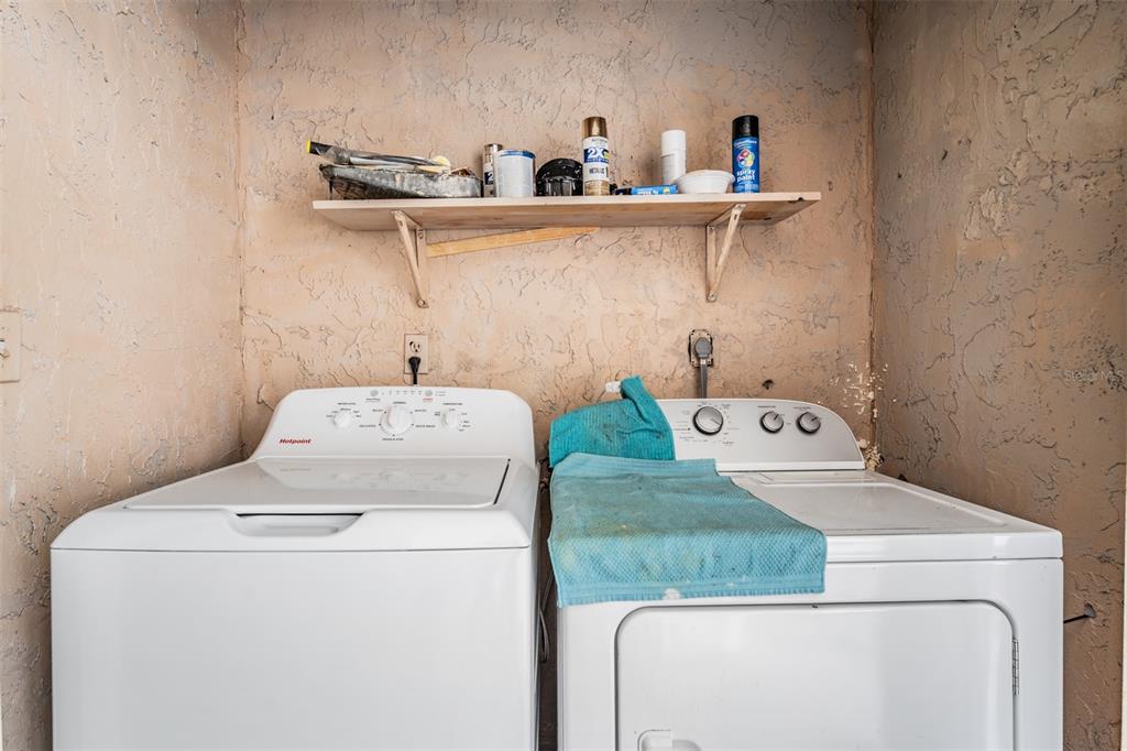 A laundry room features a white washer and dryer beneath a simple wooden wall shelf — 7210 N MANHATTAN AVENUE UNIT 1913, TAMPA, FL
