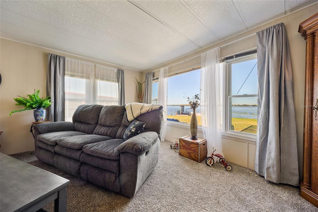 A living room with a gray sofa, carpeted floors, and large windows overlooking a lake — 121 BONNIE DRIVE, AUBURNDALE, FL