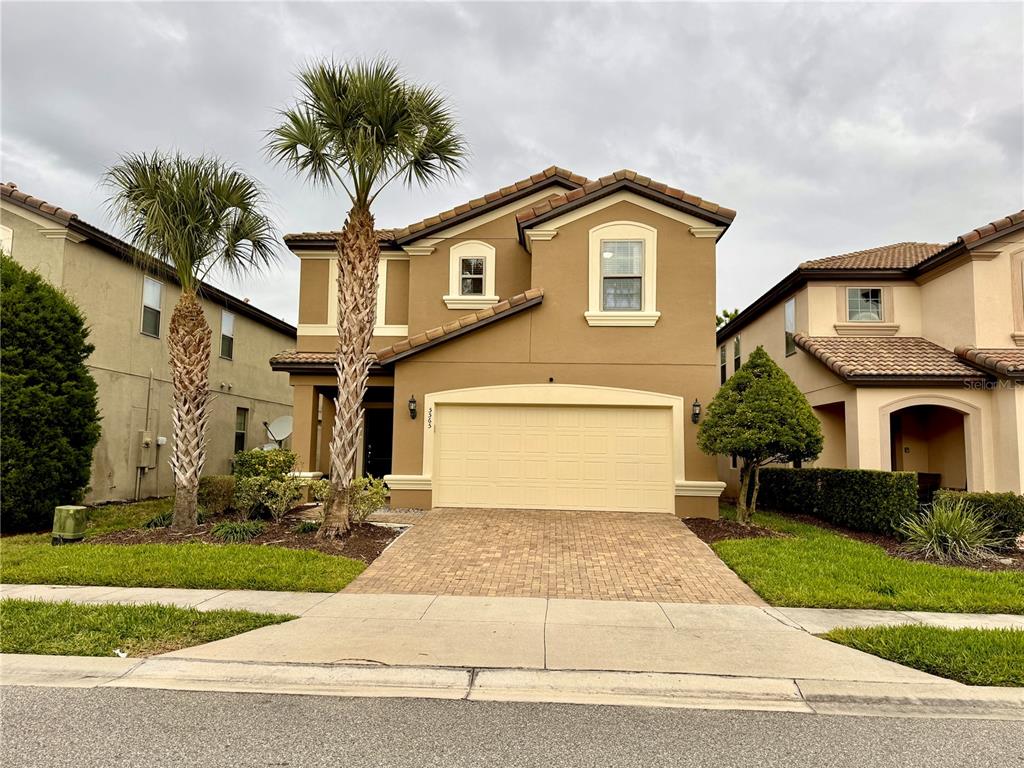 A two-story tan stucco house features a two-car garage, a paver driveway, and two palm trees — 5565 MISTY OAK CIRCLE, DAVENPORT, FL