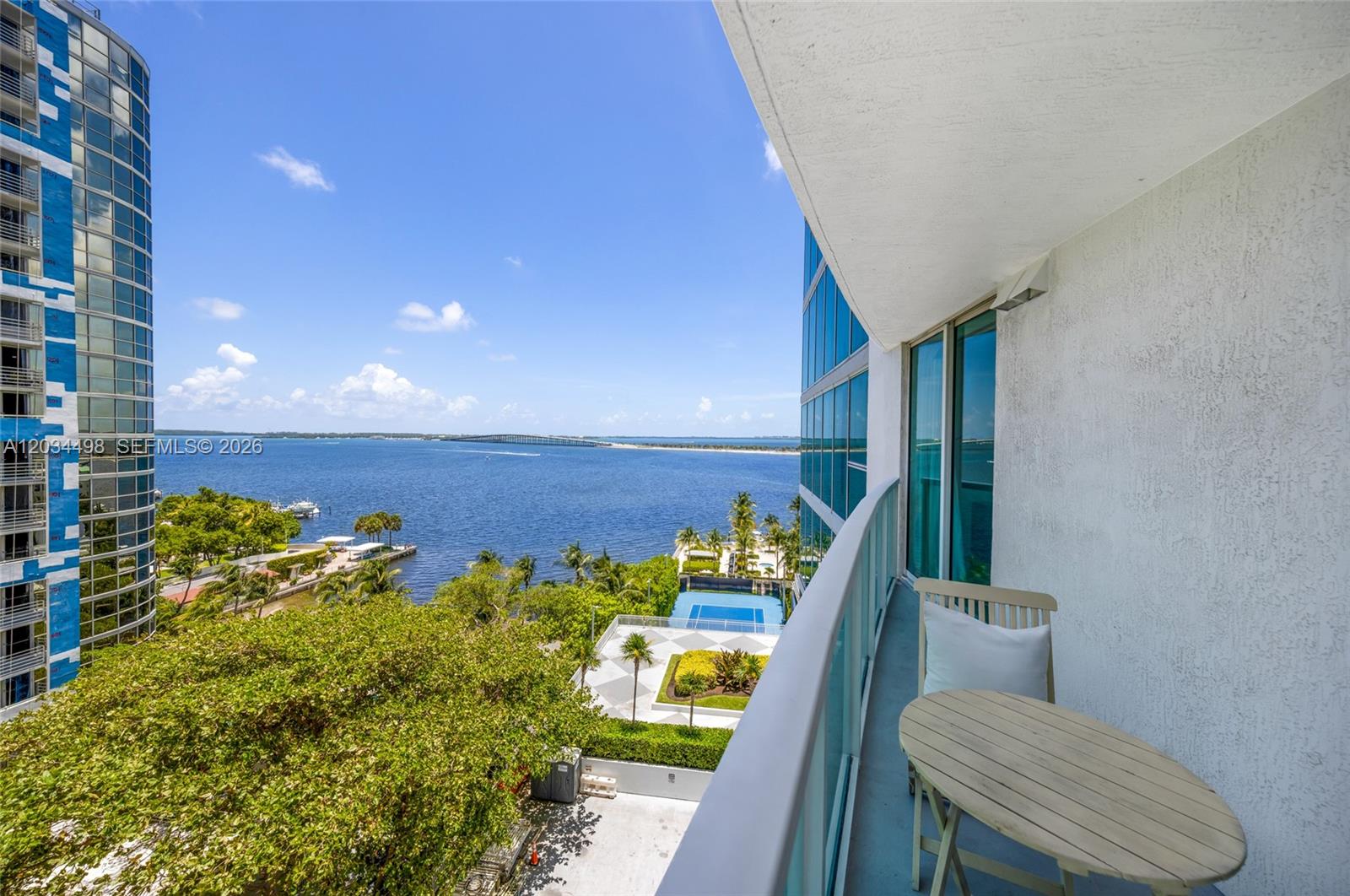 A balcony with a small table and chairs overlooks a bay, trees, and a pool — 2101 Brickell Ave # 605, Miami FL 33129, MIAMI, FL