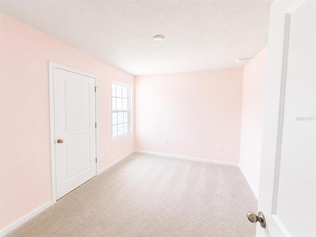 An empty bedroom features light pink walls, beige carpeting, a white door, and a window — 2008 EDISON POINTE BOULEVARD, SOUTH DAYTONA, FL