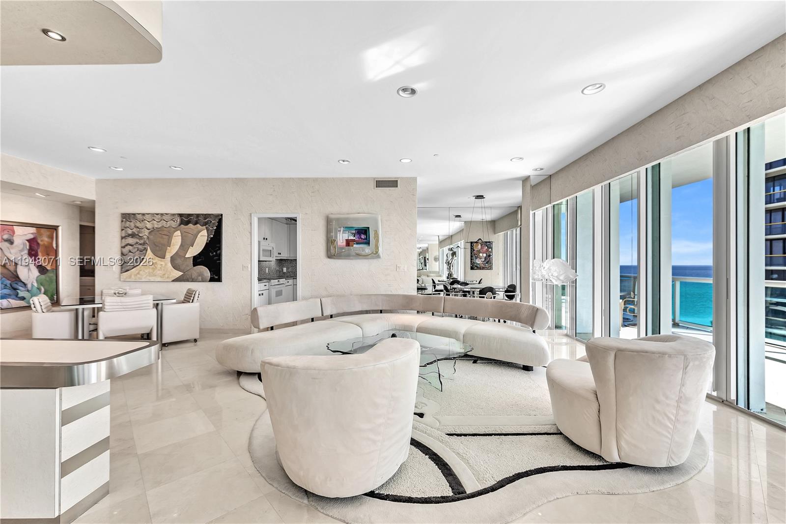 A modern living room features a curved white sofa, plush armchairs, and floor-to-ceiling windows — 10101 Collins Ave, Bal Harbour FL 33154, BAL HARBOUR, FL