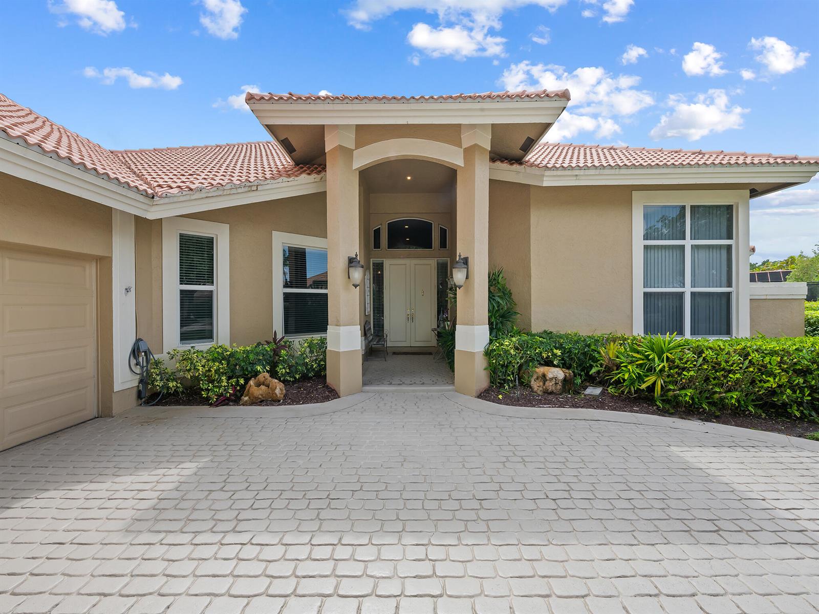A beige single-story house features a paved driveway, a tiled roof, and lush landscaping — 1207 Manor Drive S, Weston FL 33326, WESTON, FL