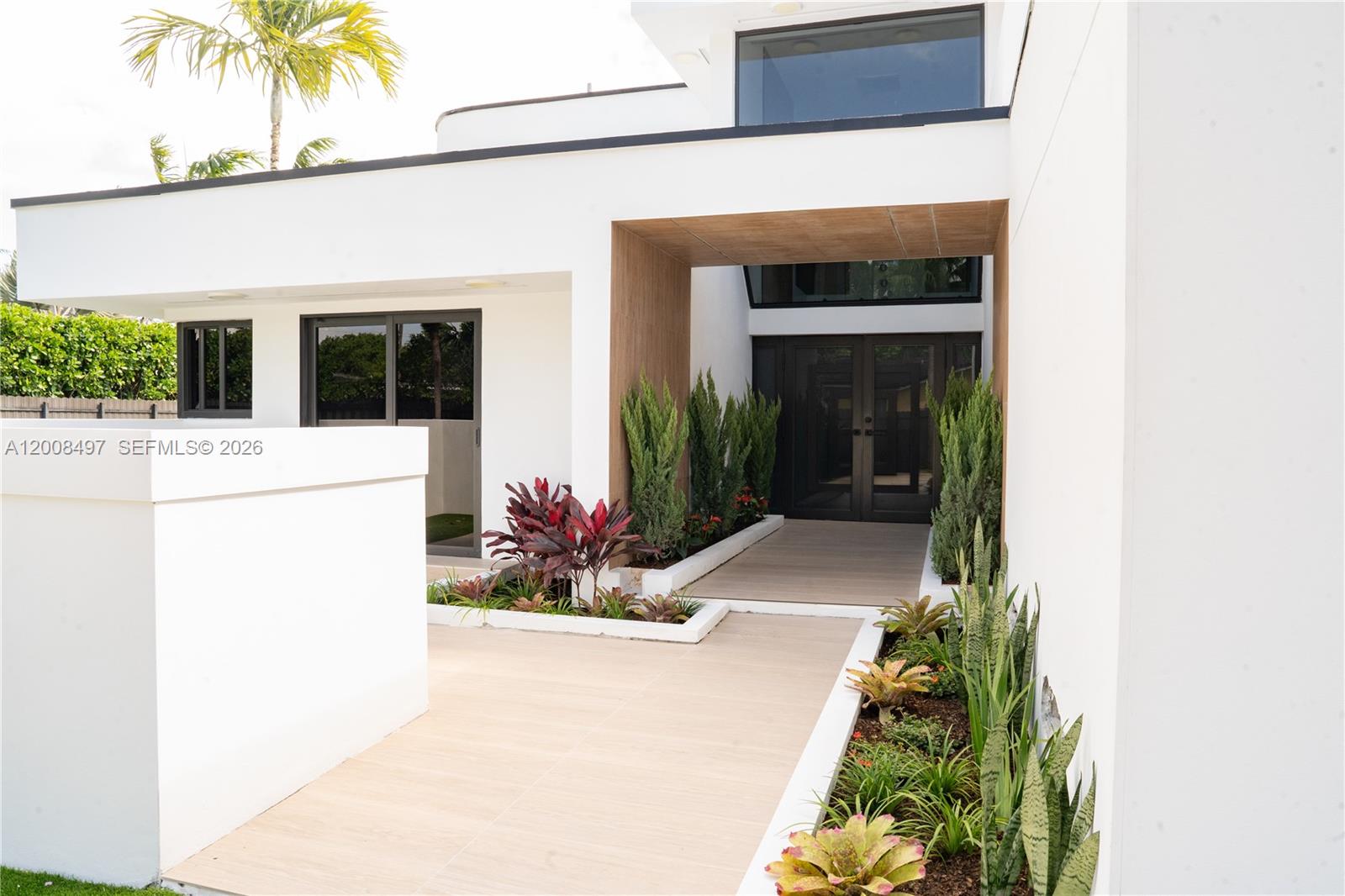The exterior of a modern white house features a wood-accented entryway and landscaped garden beds — 10475 SW 72nd Ave, Pinecrest FL 33156, PINECREST, FL