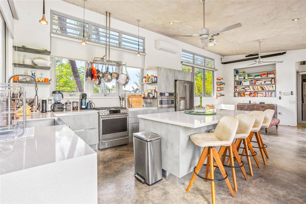 A modern kitchen features concrete floors, a large island with bar stools, and stainless appliances — PR 413 SOLAR C BO. PUNTAS, RINCON, PR