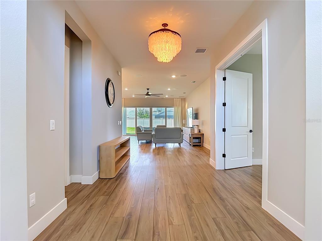 A hallway with wood-look flooring leads into a living room with a chandelier and furniture — 8891 DRUMMER PLANK DRIVE, WESLEY CHAPEL, FL