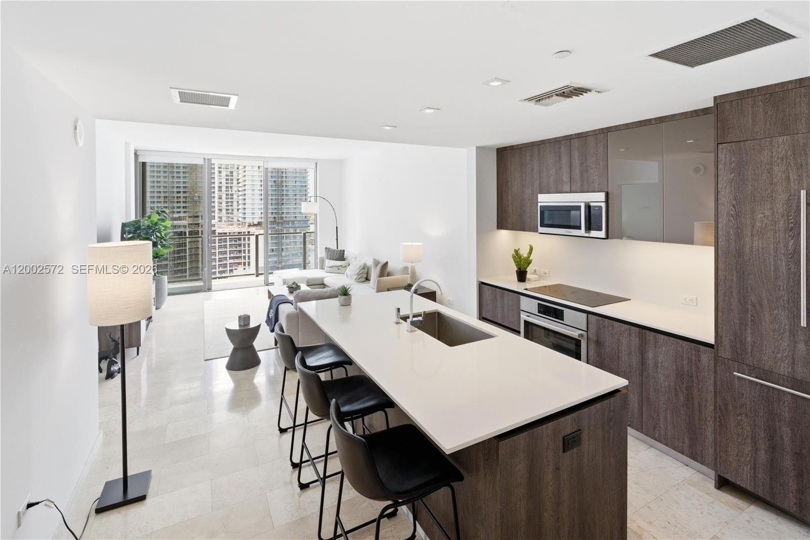 A modern kitchen with dark wood cabinetry and a white island overlooks a living area — 68 SE 6th St # 1106, Miami FL 33131, MIAMI, FL