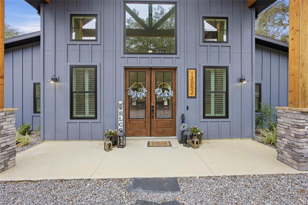 The front exterior of a blue board-and-batten house features double wood doors and stone columns — 12052 SE 101ST COURT ROAD, BELLEVIEW, FL