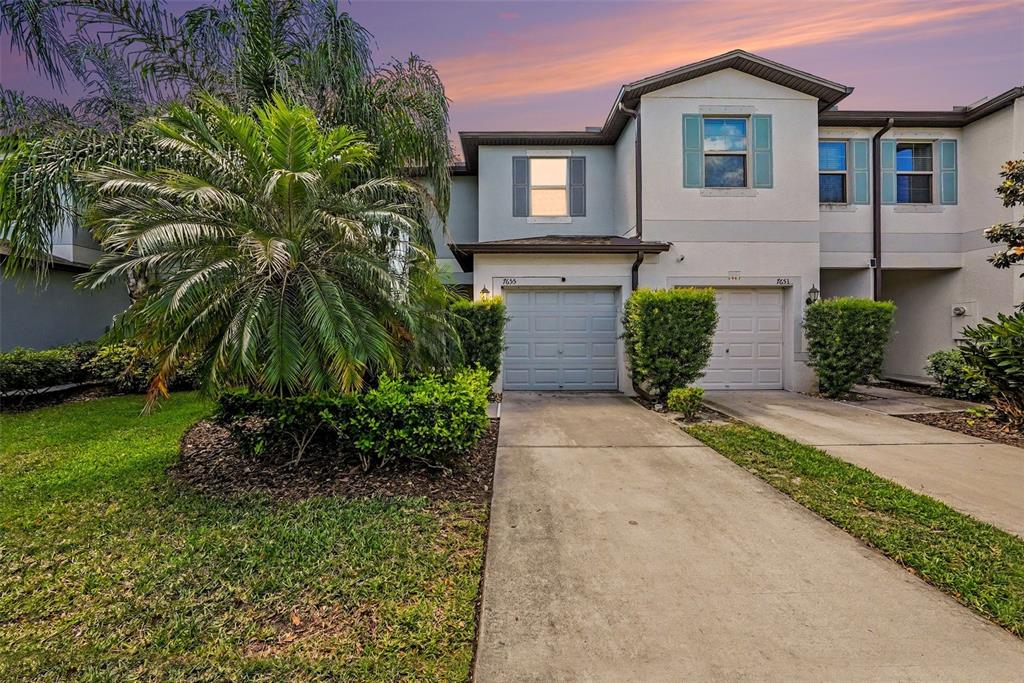 A two-story townhouse exterior features two garage doors, light-colored siding, and a palm tree — 7655 GINGER LILY COURT, TAMPA, FL