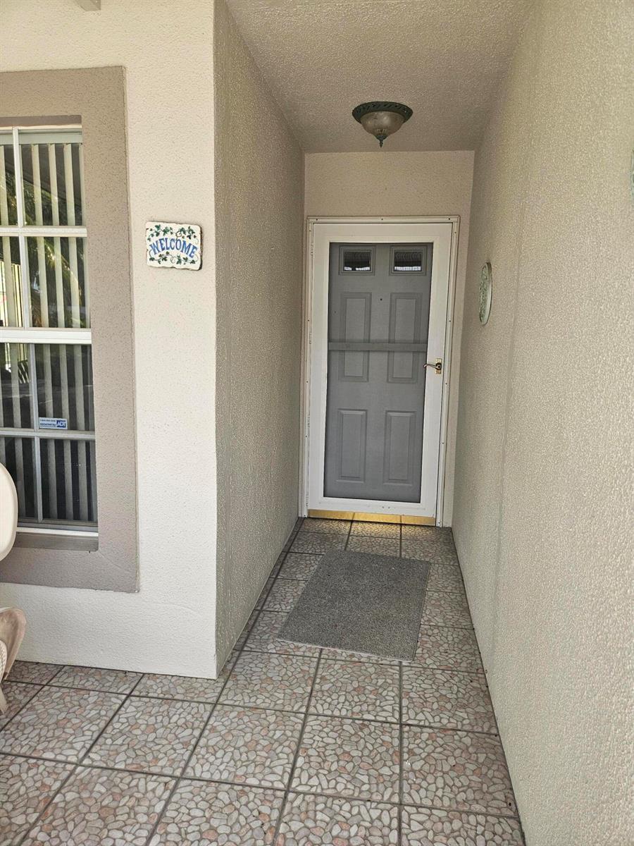 A covered entryway features a tiled floor, a grey storm door, and a welcome sign — 18999 Stewart Circle # 8, Boca Raton FL 33496, BOCA RATON, FL