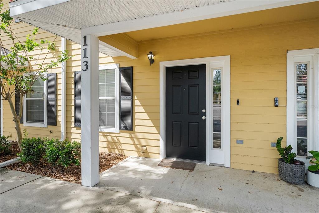 A yellow-sided house exterior features a black front door, a concrete porch, and address numbers — 2927 SW 35TH PLACE UNIT 113, GAINESVILLE, FL