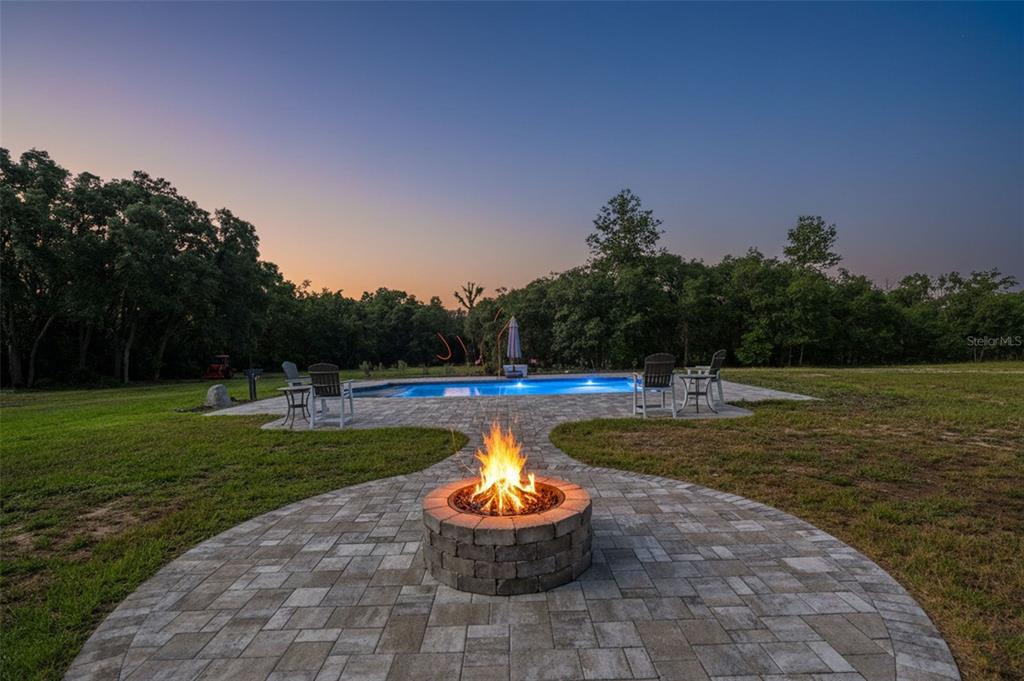 A stone fire pit sits on a paved patio in front of a backyard swimming pool — 10030 ORCHARD WAY, SPRING HILL, FL