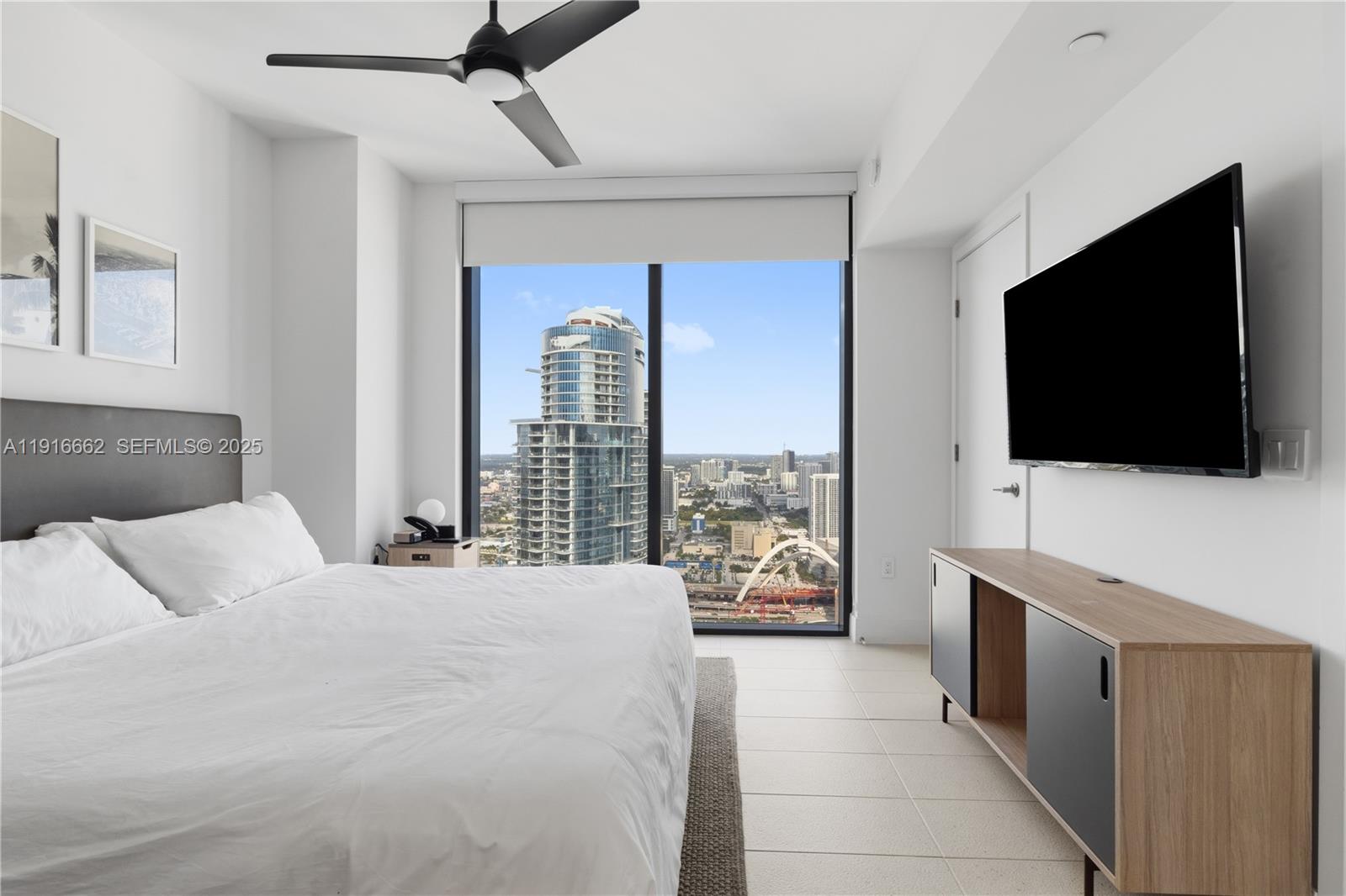 A modern bedroom features a bed, a wall-mounted television, and floor-to-ceiling windows overlooking a city — 159 NE 6th St # 4713, Miami FL 33132, MIAMI, FL