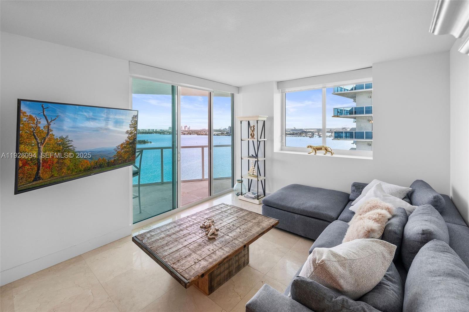 A living room features a grey sectional sofa, wooden coffee table, and floor-to-ceiling water views — 665 NE 25th St # 1801, Miami FL 33137, MIAMI, FL