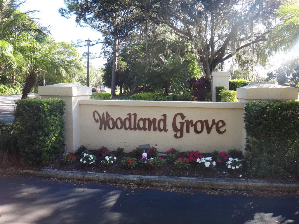 A beige stucco monument sign for Woodland Grove sits in front of trees and landscaping — 4493 LONGMEADOW UNIT 76, SARASOTA, FL