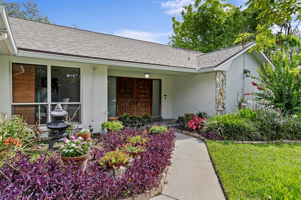 A single-story house exterior features a stone accent pillar, double wooden doors, and purple landscaping — 6314 E 112TH AVENUE, TAMPA, FL