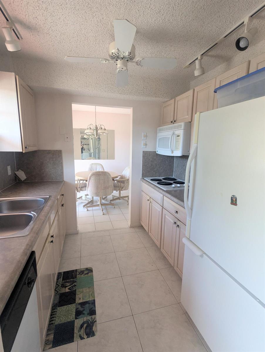 A galley kitchen with light wood cabinets, white appliances, and tiled floors leads to a dining area — 2 Golfs Edge # D, West Palm Beach FL 33417, WEST PALM BEACH, FL