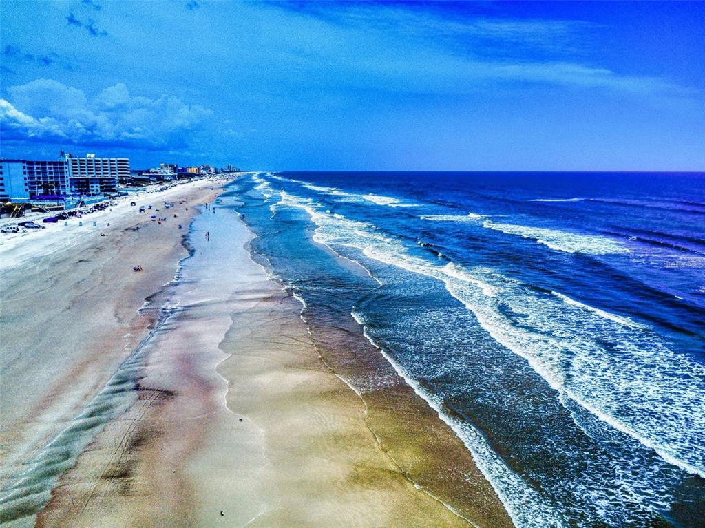 An aerial view of a sandy beach with ocean waves and coastal buildings in the distance — 1913 HILL STREET UNIT 1, NEW SMYRNA BEACH, FL