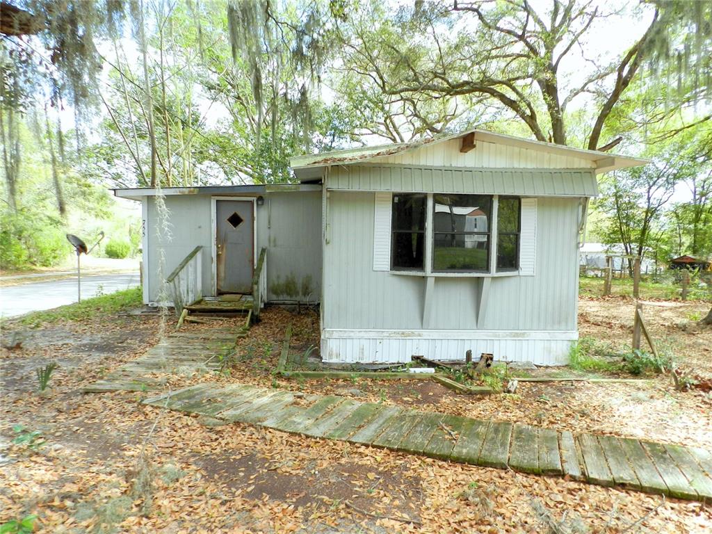 A weathered, light-colored mobile home features a bay window, a wooden walkway, and surrounding trees — 755 SW JASMINE AVENUE, KEYSTONE HEIGHTS, FL