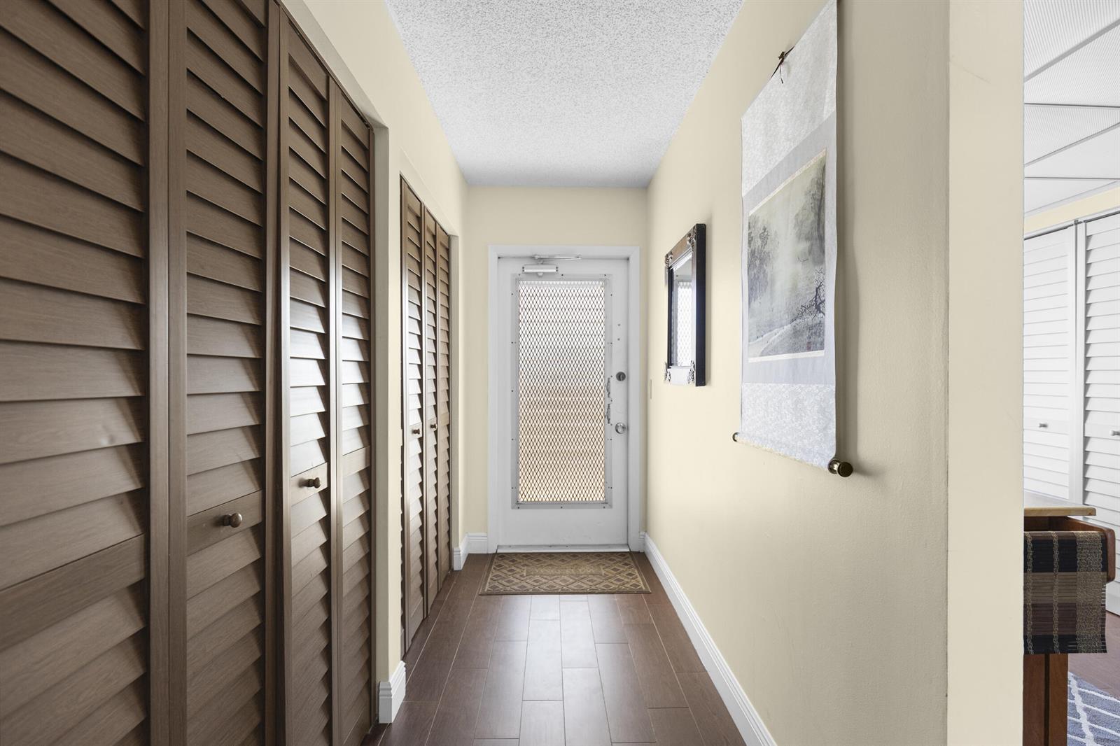 A hallway features wood-look flooring, louvered closet doors, a wall hanging, and a white entry door — 14671 Bonaire Boulevard # 404, Delray Beach FL 33446, DELRAY BEACH, FL