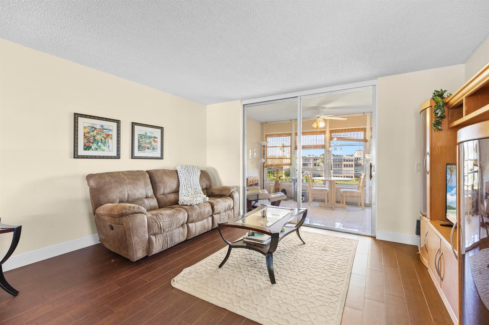 A living room with wood flooring, a brown sofa, a glass coffee table, and a lanai — 14671 Bonaire Boulevard # 404, Delray Beach FL 33446, DELRAY BEACH, FL