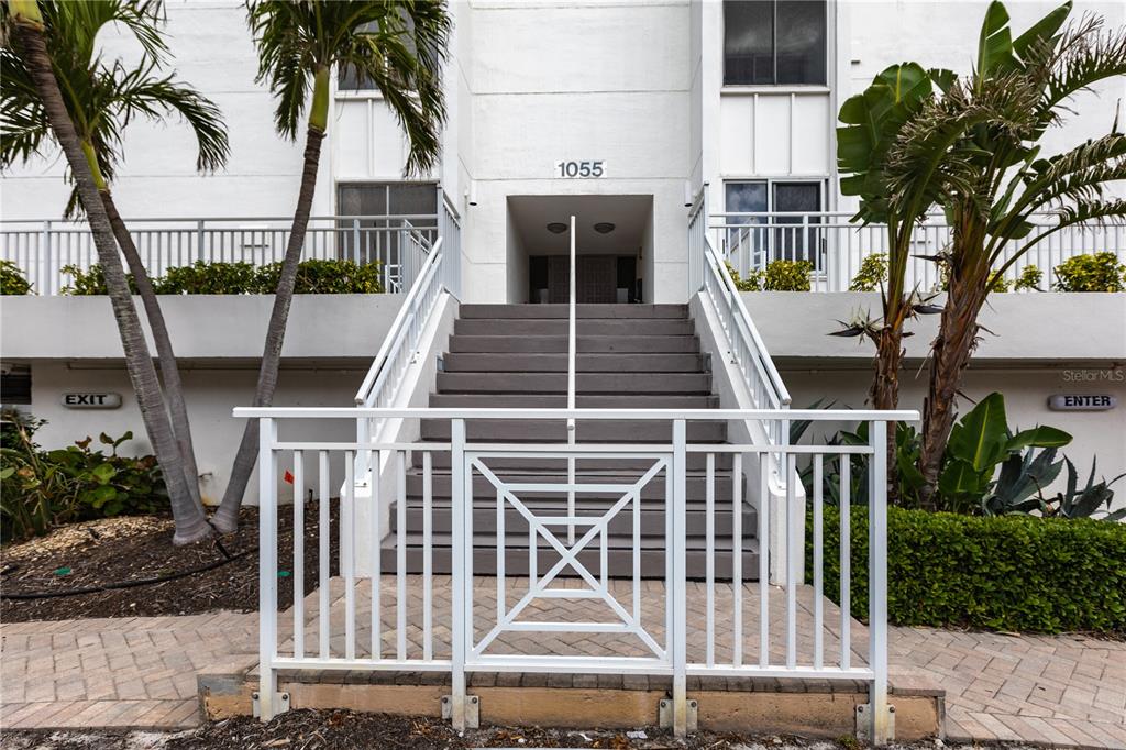 A white building entrance features a central staircase with a metal railing and palm trees — 1055 W PEPPERTREE DRIVE UNIT 703AA, SARASOTA, FL
