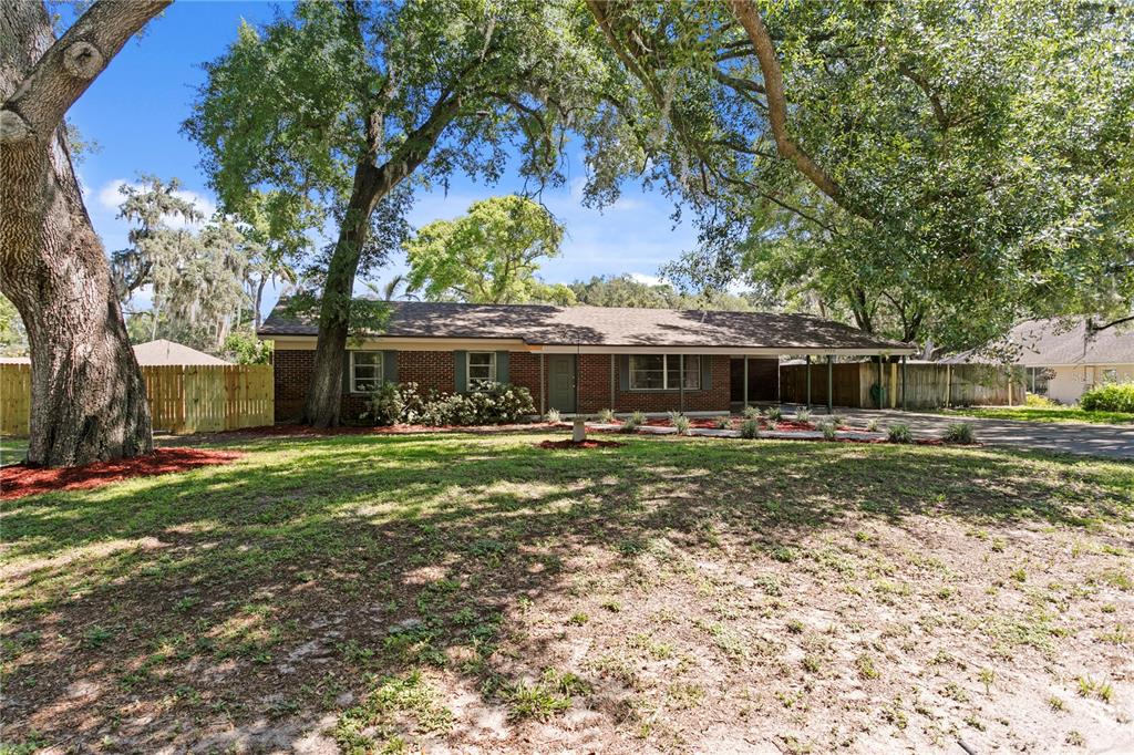 A single-story brick house with a carport sits on a grassy lot under large trees — 6928 GREENHILL PLACE, TAMPA, FL